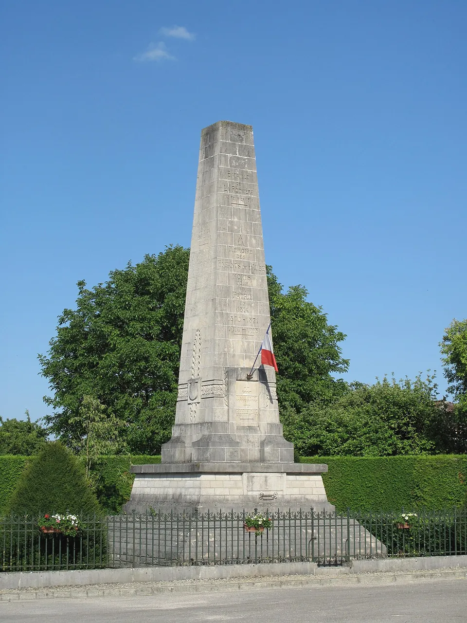 Stone memorial standing beside a track, enclosed by a low railing, in the open farmland of the Aube