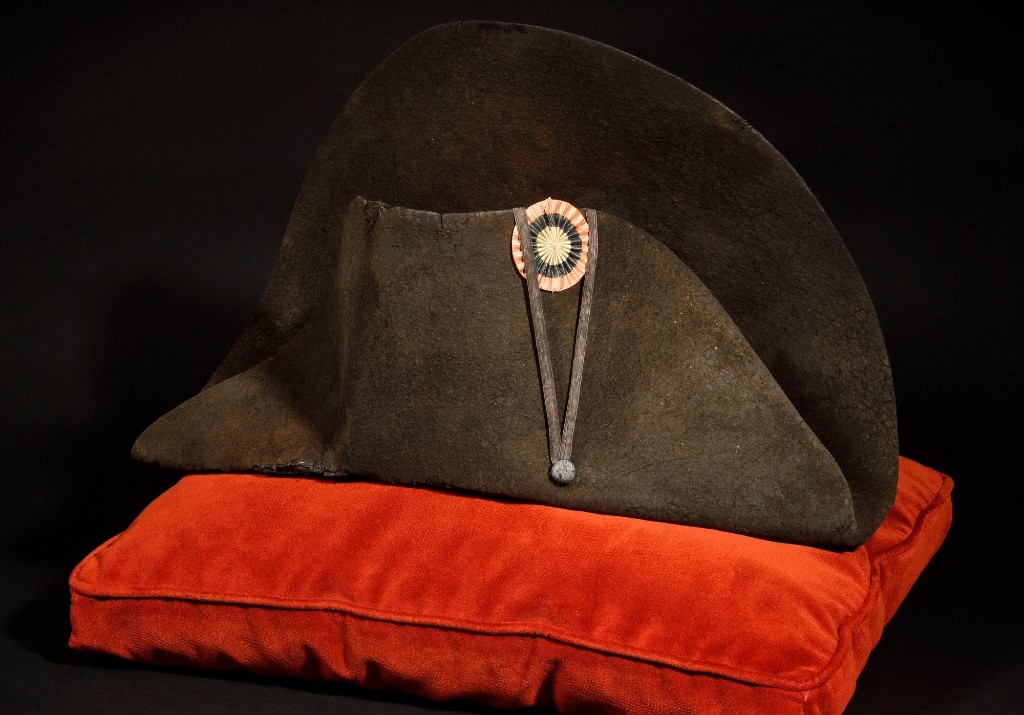 Bicorn Napoleons I. — schwarzer Filzhut quer getragen mit Trikolorkokarde, auf einem roten Samtkissen, ikonisches Emblem des Ersten Kaiserreichs