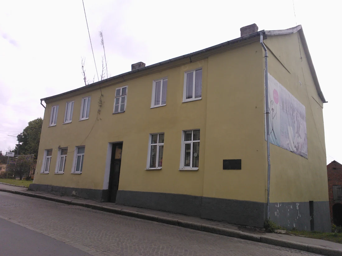 Two-storey house with pale yellow walls along a paved street, a commemorative plaque on its gable