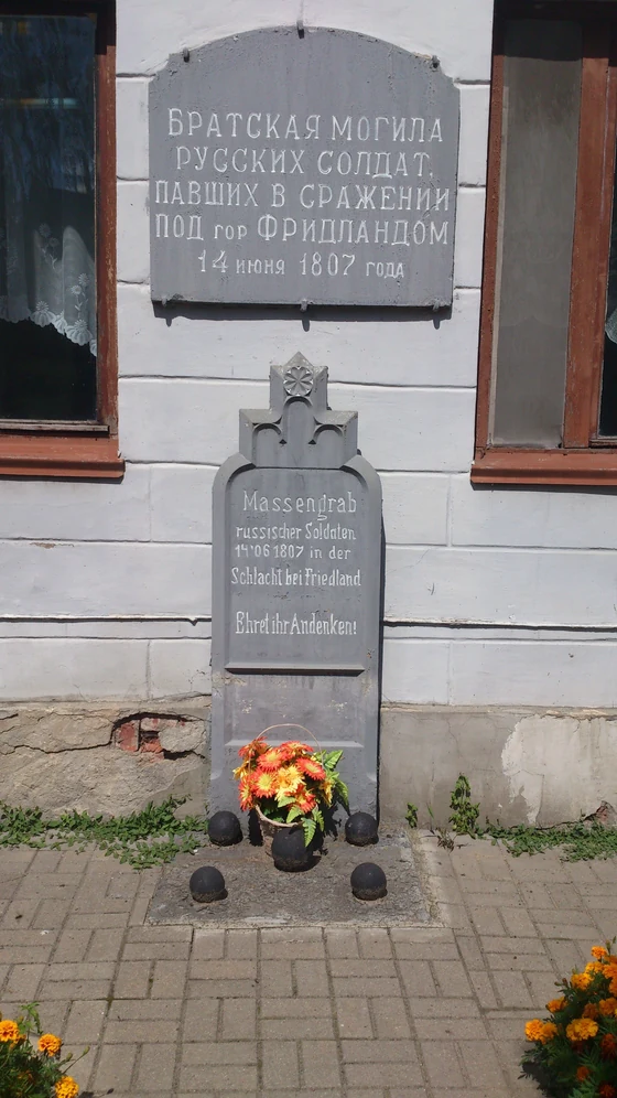 Pale façade bearing two stone plaques engraved in Cyrillic and German, with flowers laid at the foot