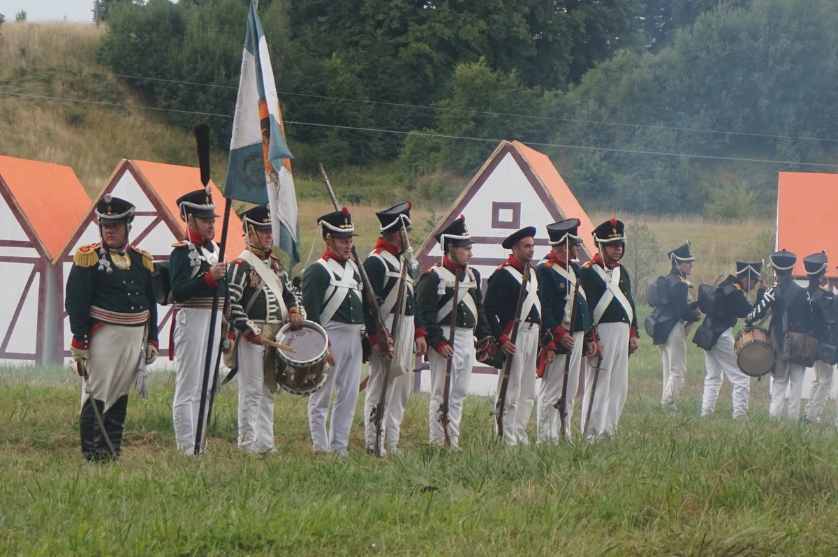 Re-enactors in white and green Russian uniforms lined up under a flag in front of tents, on a meadow