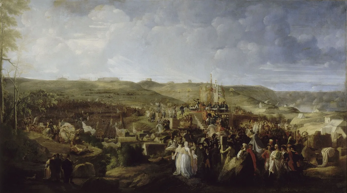 Vaste paysage de côte vallonnée : au centre, une estrade surmontée de trophées où se tient l'Empereur, entourée d'une foule immense de soldats en rangs, de cavaliers et de spectateurs, sous un grand ciel nuageux