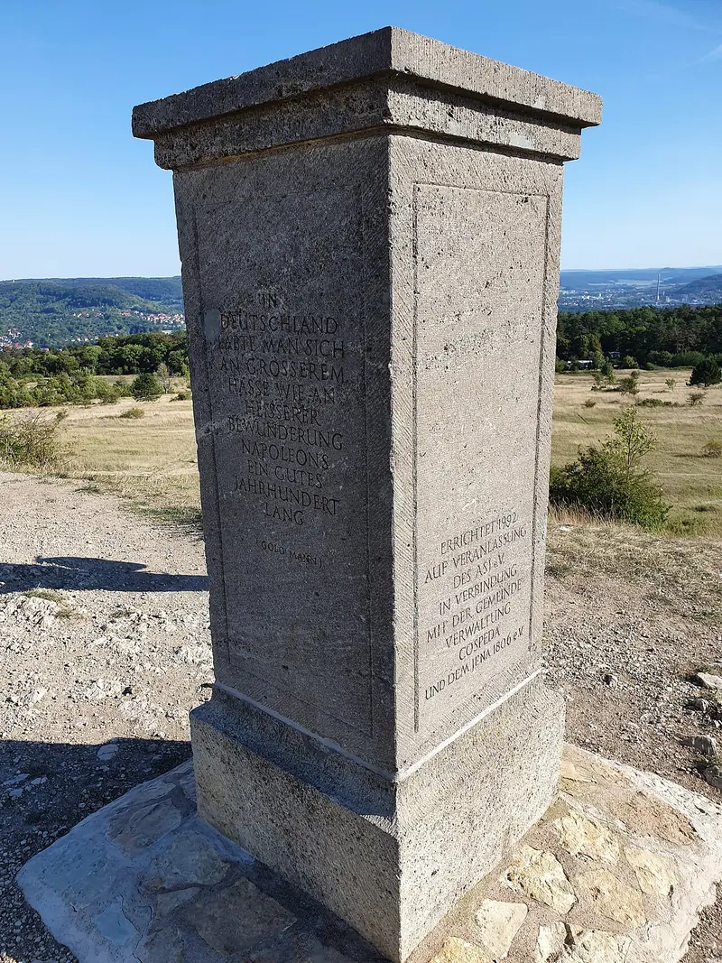 Estela de piedra cuadrada en una altura pelada, con el valle del Saale y la ciudad de Jena a lo lejos
