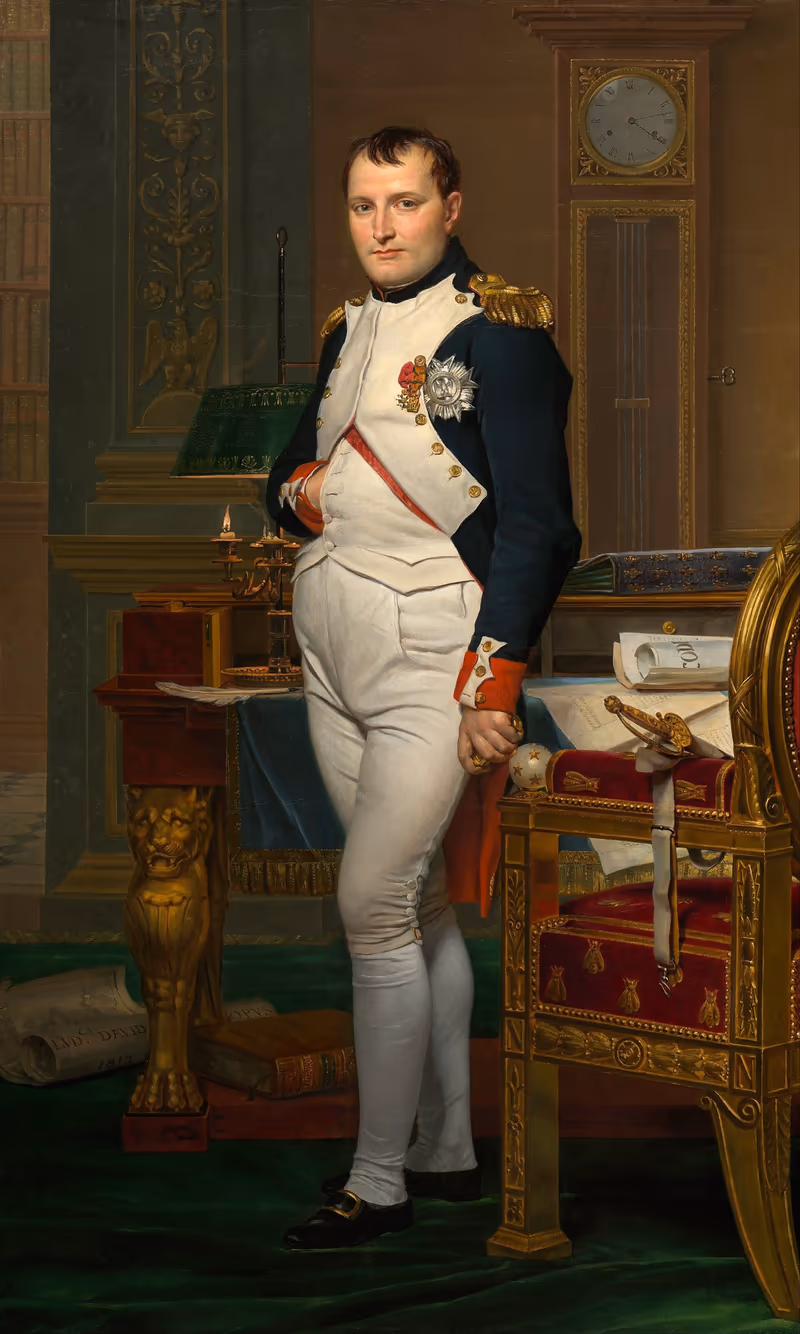 Napoléon en uniforme blanc et rouge, debout dans son cabinet de travail des Tuileries, la main droite glissée dans son gilet, près d'un bureau couvert de papiers