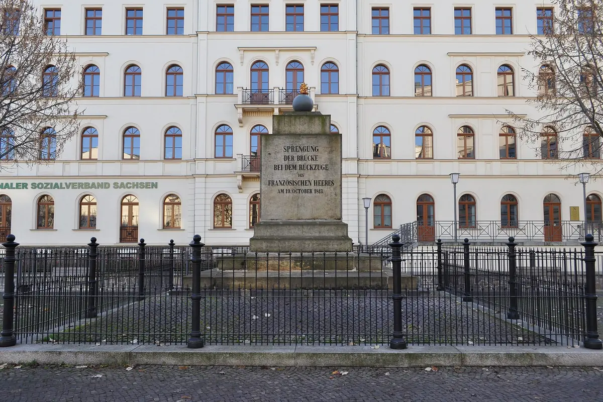 Monumento di pietra a gradoni sormontato da una sfera di ghisa, circondato da una cancellata, davanti a un grande edificio chiaro