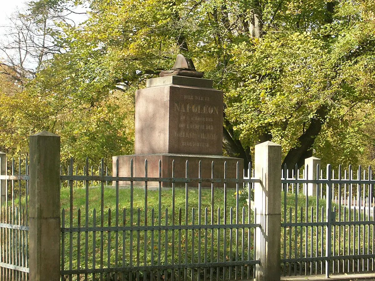 Blocco di granito rosato con il nome di Napoleone, sormontato da un cappello scolpito, circondato da una cancellata in un parco