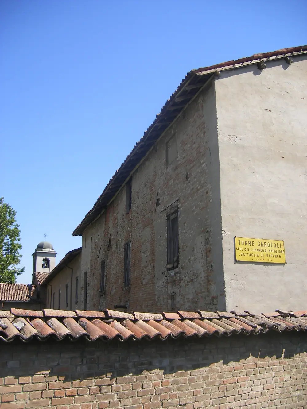 Long brick and stone building with a tiled roof and a bell turret, behind a wall