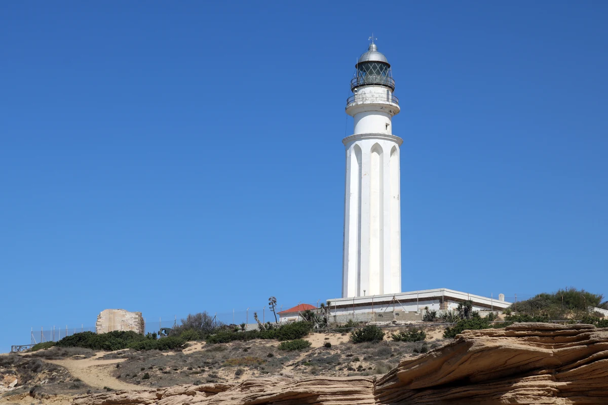 White lighthouse on a promontory of ochre rocks, beside a ruined tower, under a blue sky