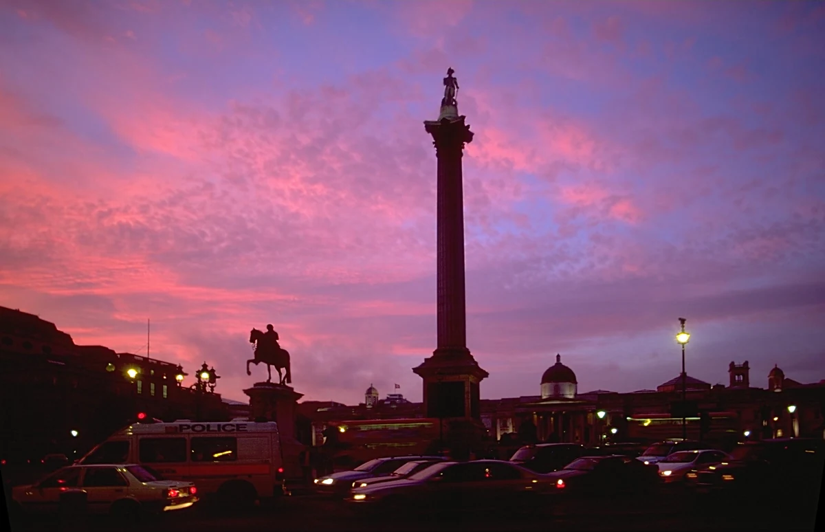 Square at dusk under a pink and violet sky: a tall column stands out, surrounded by street lamps and an equestrian statue