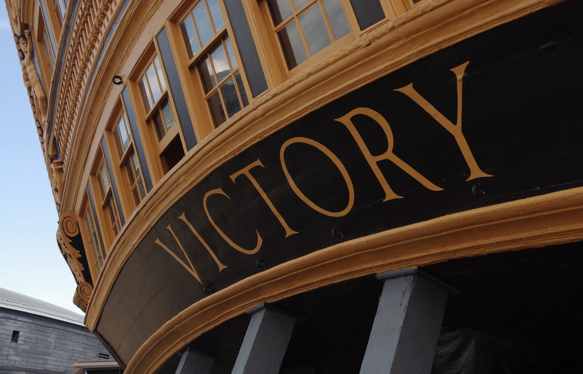 Curved stern of an old ship painted black and ochre, with its gallery windows and the name VICTORY in gilt letters