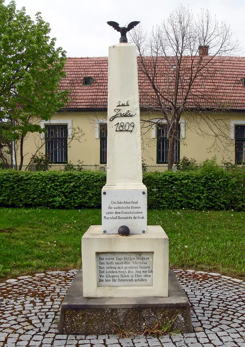 Obélisque blanc surmonté d'un aigle noir, portant la date des 5 et 6 juillet 1809 et un boulet, devant une maison de village