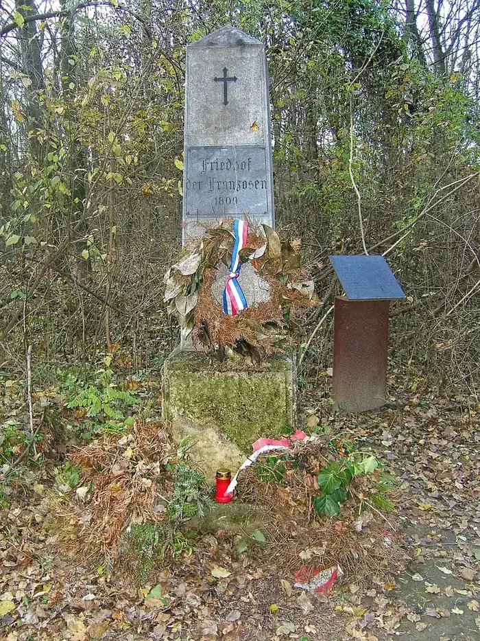 Stèle de pierre gravée d'une croix et de l'inscription « Friedhof der Franzosen 1809 », ornée d'une couronne à ruban tricolore, dans un sous-bois